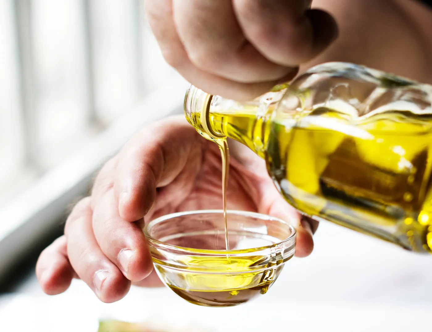 Olive oil poured from a bottle into a small glass bowl held in a hand.
